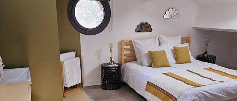 A bedroom with a large bed, a round window, a bedside table, and a nightstand.