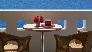 En balkon med fletstole, et rundt bord med en vase blomster, og en rød genstand på bordet. I baggrunden ses udsigt over havet.