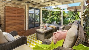 Ein überdachter Patio-Bereich mit Rattanmöbeln, einem gelb-weißen Zickzack-Teppich und Blick ins Grüne.