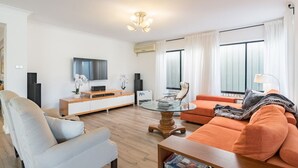 Ein modernes Wohnzimmer mit Sofa, Sessel und Couchtisch. Ein Flachbildfernseher ist an der Wand angebracht, ein kleiner Beistelltisch mit einer Pflanze steht daneben, und ein Fenster mit Vorhängen ist zu sehen.
