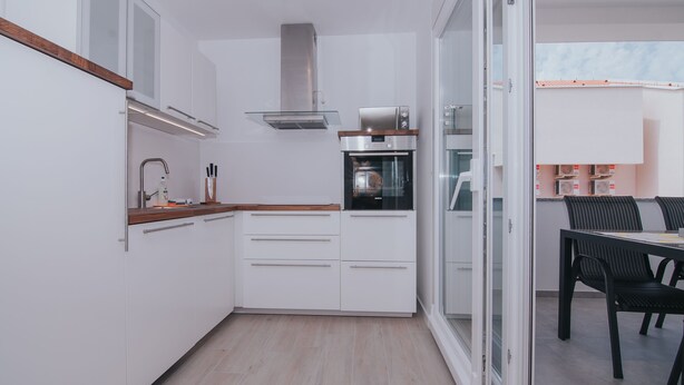 Una cocina moderna con armarios blancos, horno empotrado y comedor con mesa y sillas.