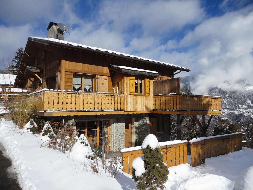 A wooden chalet with a balcony and a stone foundation, surrounded by snow and mountains.