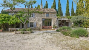 Une maison en pierre avec un toit en tuiles, une allée gravillonnée et un jardin où poussent différentes plantes et arbres.