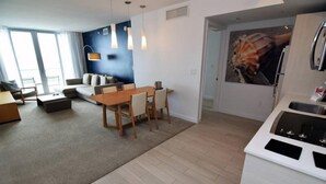 A modern living room with a sofa, coffee table, and dining area. There is a kitchen with a stove and a large window.