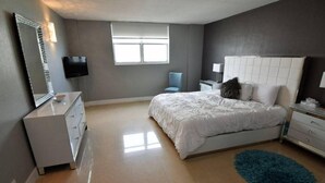 Un dormitorio con cama, cómoda, televisor, silla y ventana.