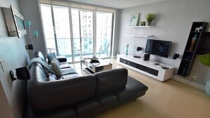 Una sala moderna con un sofá gris oscuro, un televisor de pantalla plana sobre un mueble blanco y un balcón con ventanas amplias.