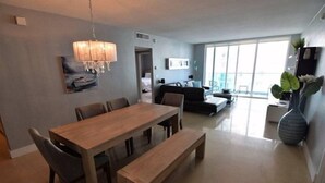 Una sala moderna con una mesa de comedor de madera, bancos y una araña. Hay un sofá y un ventanal grande que proporciona luz natural.