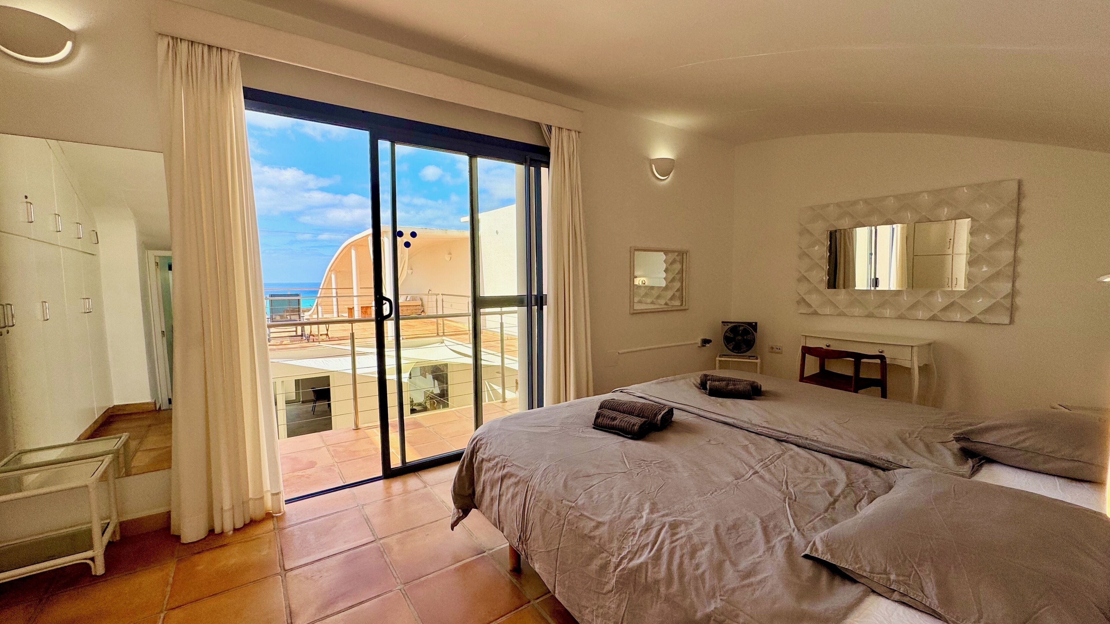 Ein Schlafzimmer mit einem großen Bett, einem Schreibtisch und Blick auf den Strand durch eine Schiebetür.
