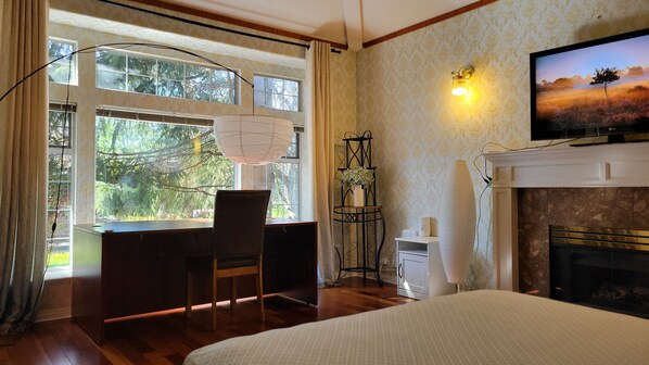 A room with a desk, chair, large window, wall-mounted TV, fireplace, and a standing lamp.