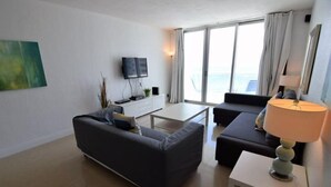 Ein modernes Wohnzimmer mit Sofa, Couchtisch und Fernseher. Ein großes Fenster mit Vorhängen und eine Lampe auf einem Beistelltisch.
