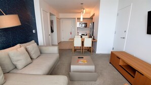 A modern living room with a sofa, ottoman, and a TV stand. There is a dining area with a table and chairs, and a kitchen in the background. - Ocean & Bay View Residence 1 Bed 1 Bath @ Beachwalk Hallandale Beach (Hallandale Beach)