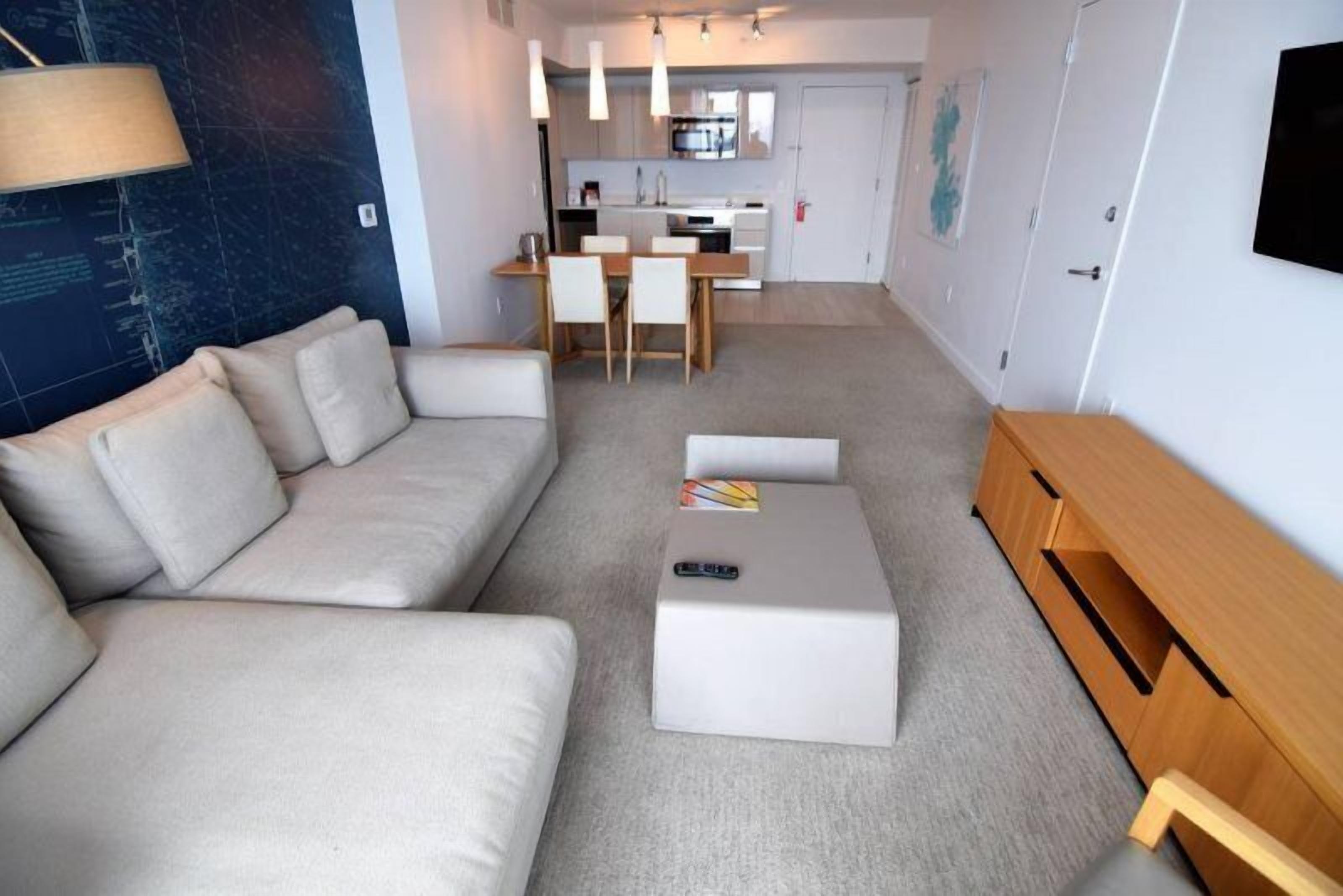 Un salon moderne avec un canapé beige, une table basse et un meuble pour la télévision. On aperçoit une cuisine à l’arrière-plan, avec une table à manger et des chaises.