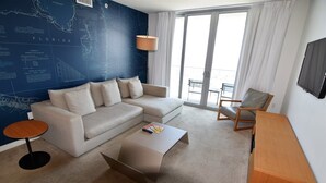 Ein modernes Wohnzimmer mit einem Ecksofa, einem kleinen runden Tisch, einem Couchtisch, einem Holznachttisch, einem an der Wand montierten Fernseher und einem großen Fenster mit Vorhängen.