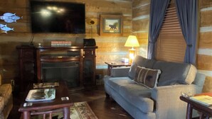 Interior acogedor de una cabaña de madera con techo de madera, ventilador de techo, televisor, chimenea, sofá, mesa de centro y lámpara.