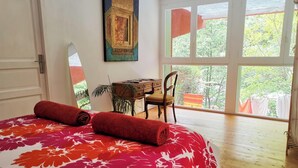A bedroom with a large bed, a desk, and a chair. There is a window with a view of trees and a balcony with clothes hanging. - Beautiful River-front Apartment with Natural Pool (Sorède)