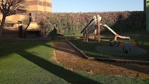 A playground with a slide and swings, surrounded by a hedge and a building in the background. - Apartment towers, wifi, pool, breakfast. (Calahorra)