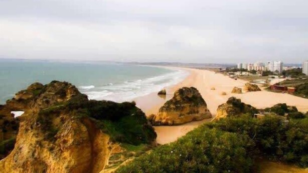 A coastal landscape with rocky outcrops, a sandy beach, and a distant cityscape. - Liiiving in Algarve | Alvor Blue Villa (Alvor)