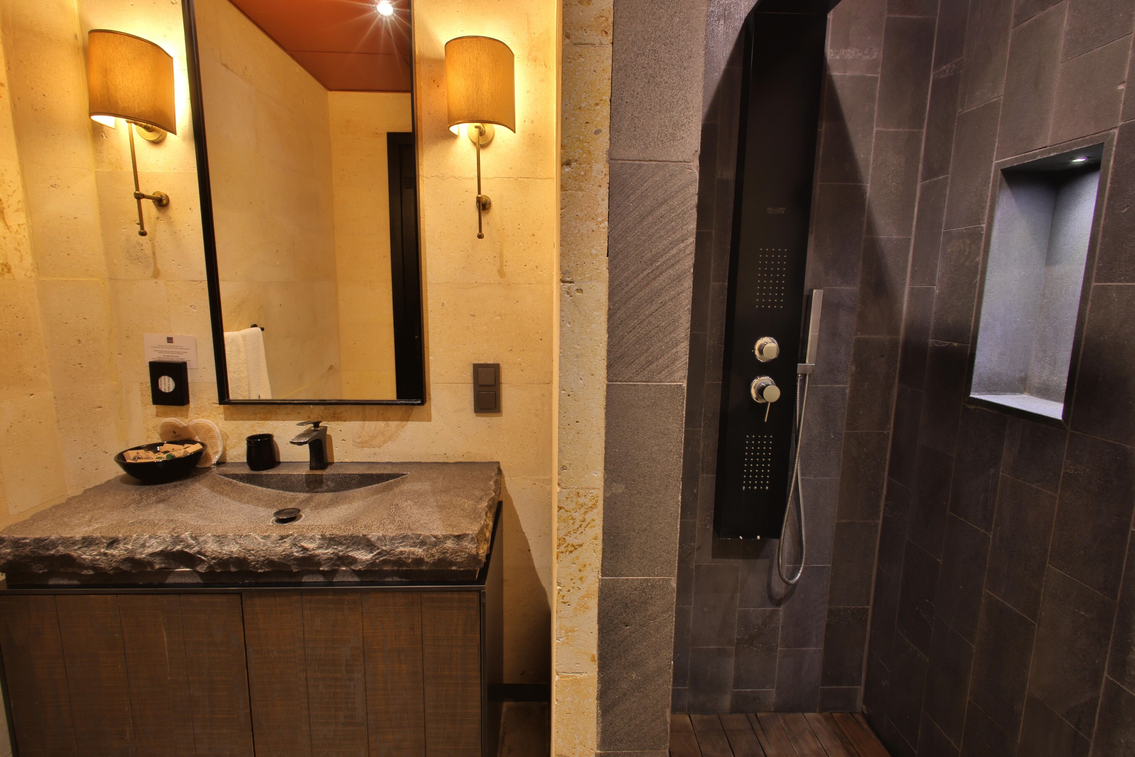 A bathroom with a granite sink, two wall-mounted lamps, a mirror, a towel dispenser, and a shower with a control panel.