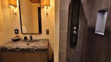 Une salle de bain avec un lavabo en granit, deux lampes fixées au mur, un miroir, un distributeur de serviettes et une douche dotée d’un panneau de commande.