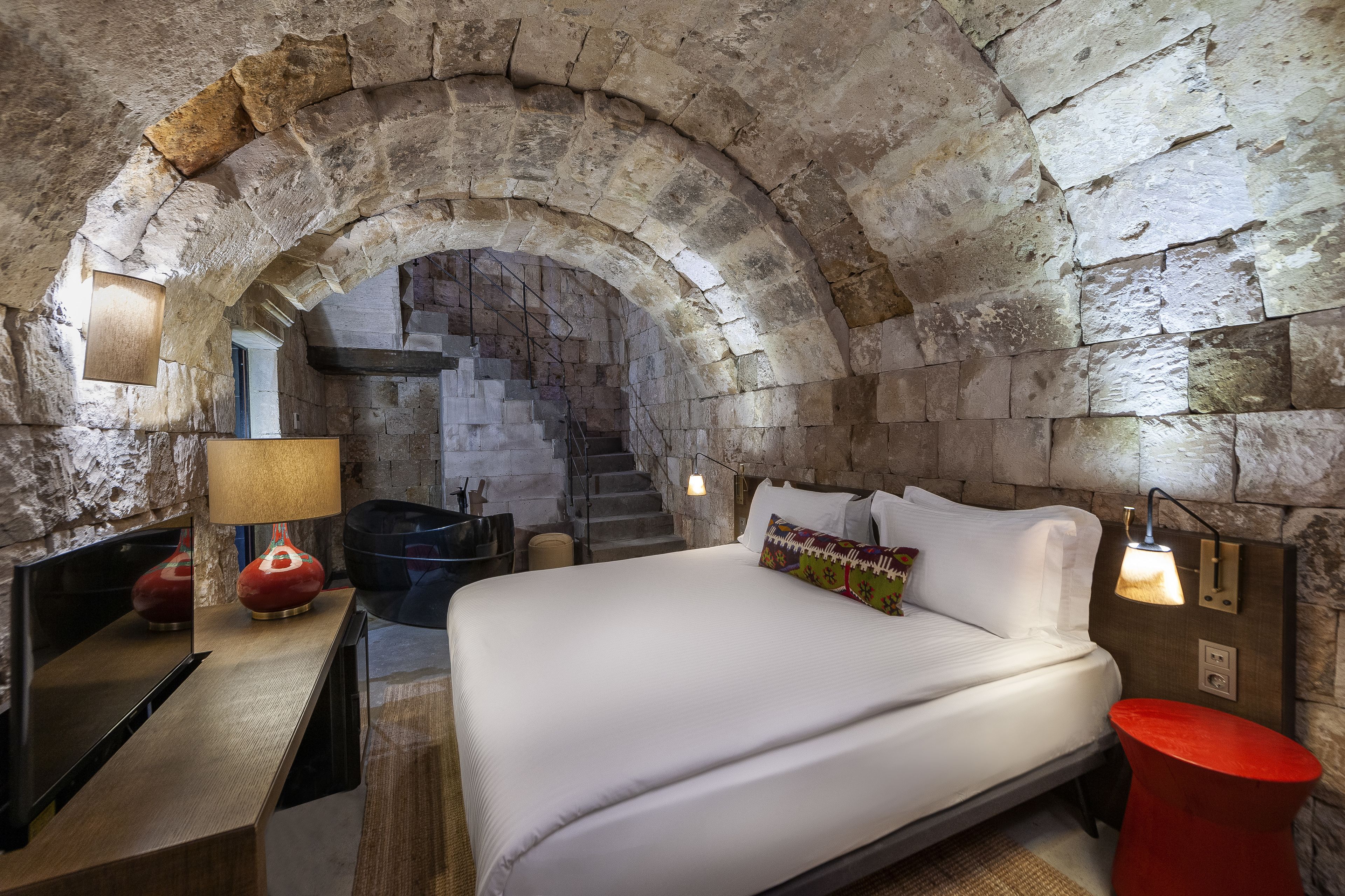 A stone-walled room with a bed, a desk, and a lamp.