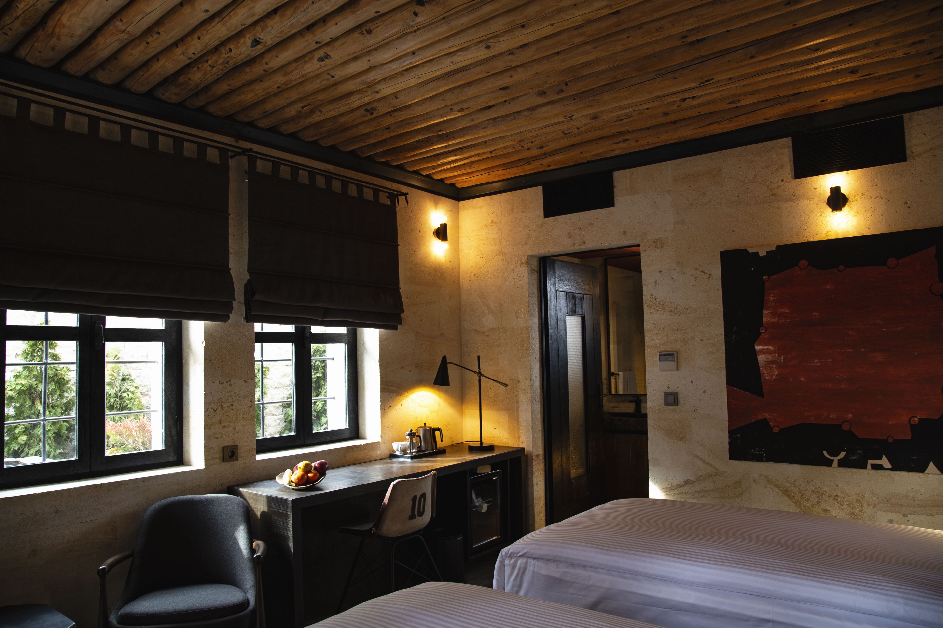 A room with two beds, a desk with a chair, a window with a view, and a wall with a red abstract painting.