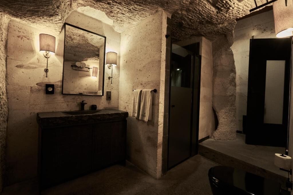A rustic bathroom with a stone wall, a wooden vanity, a mirror with wall-mounted lights, and a towel hanging on the wall.