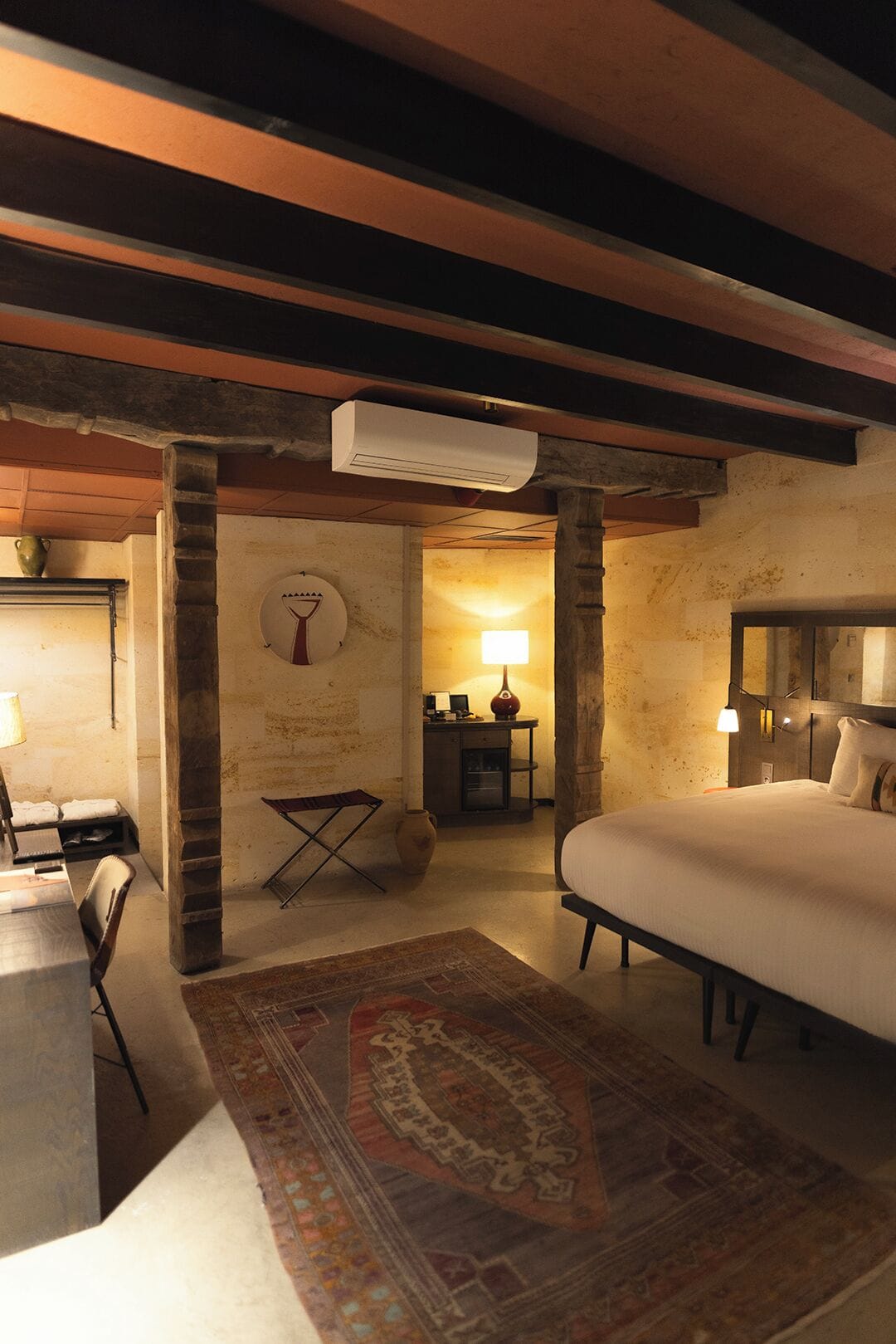 Een gezellige slaapkamer met een groot bed, een bureau met stoel, een klein tafeltje met lamp en een decoratieve vaas.