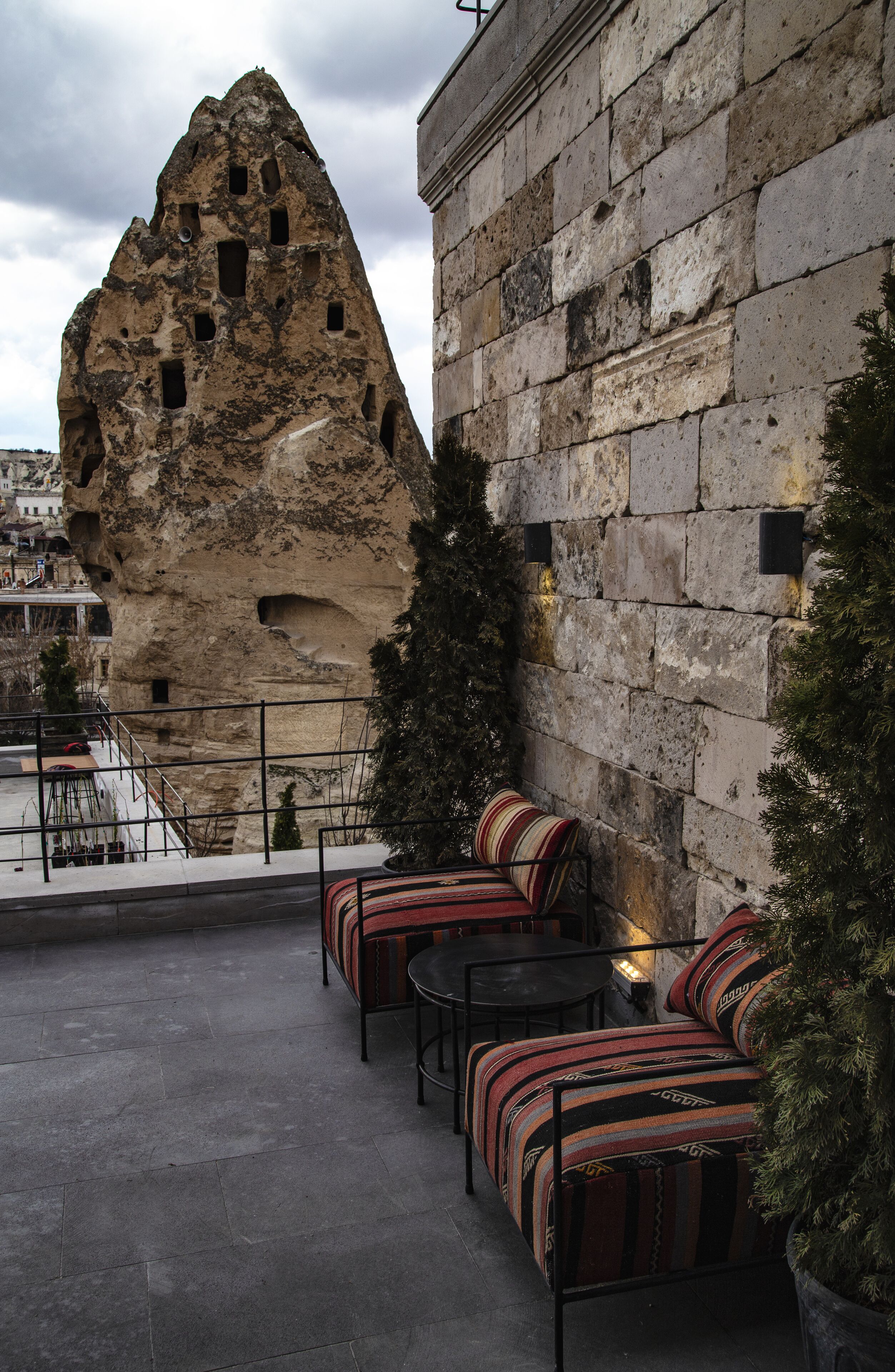 Un edificio de piedra con una gran formación rocosa, asientos exteriores con cojines a rayas y una planta en maceta.