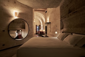 A bedroom with a large bed, a round mirror, and a view of a dining area through an arched doorway. - Carus Cappadocia (Nevsehir)