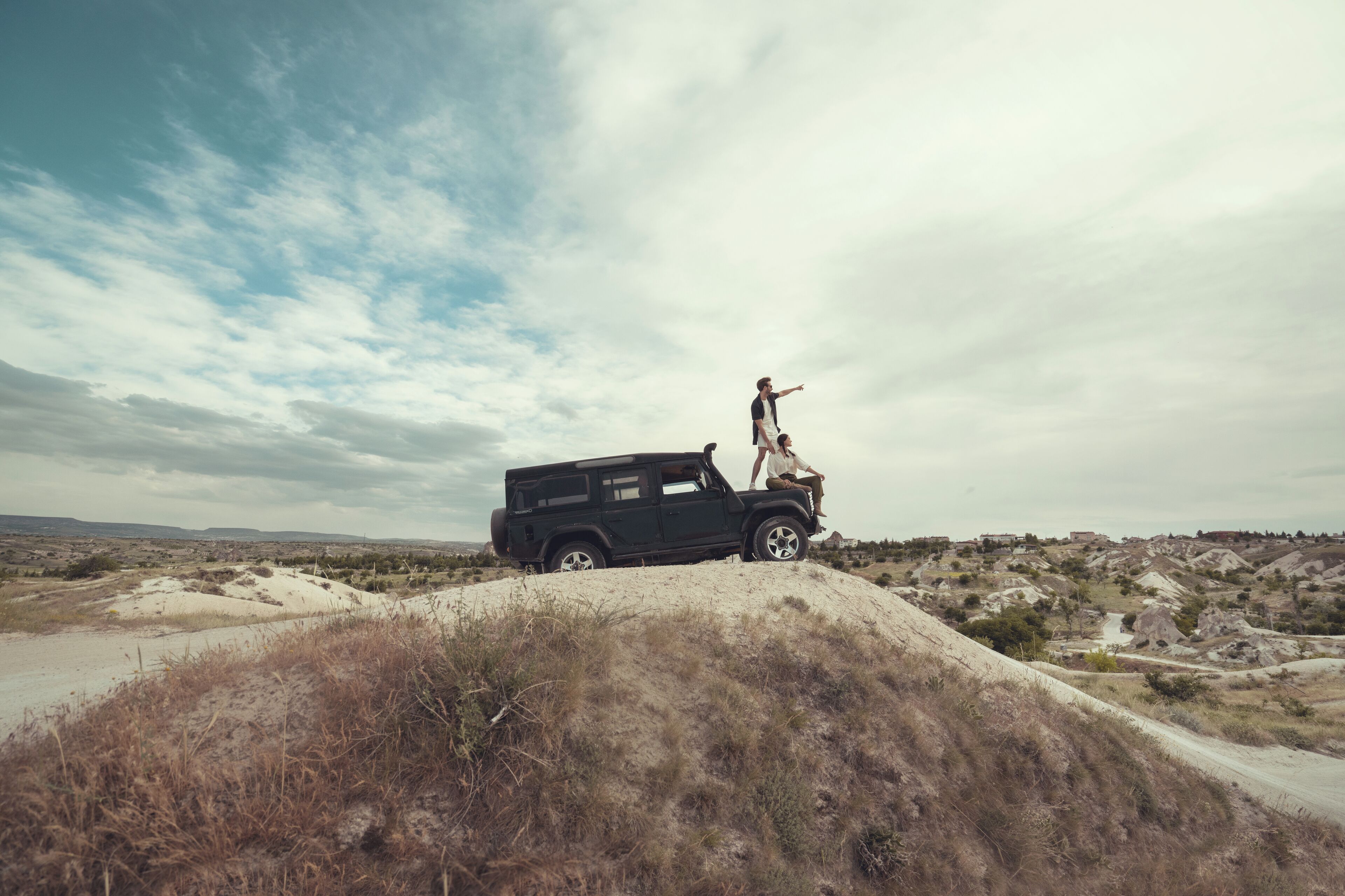 En sort SUV holder parkeret på en sandet bakke, med en person stående øverst og kigge ud over et landskab med sparsom vegetation og fjerne bygninger.