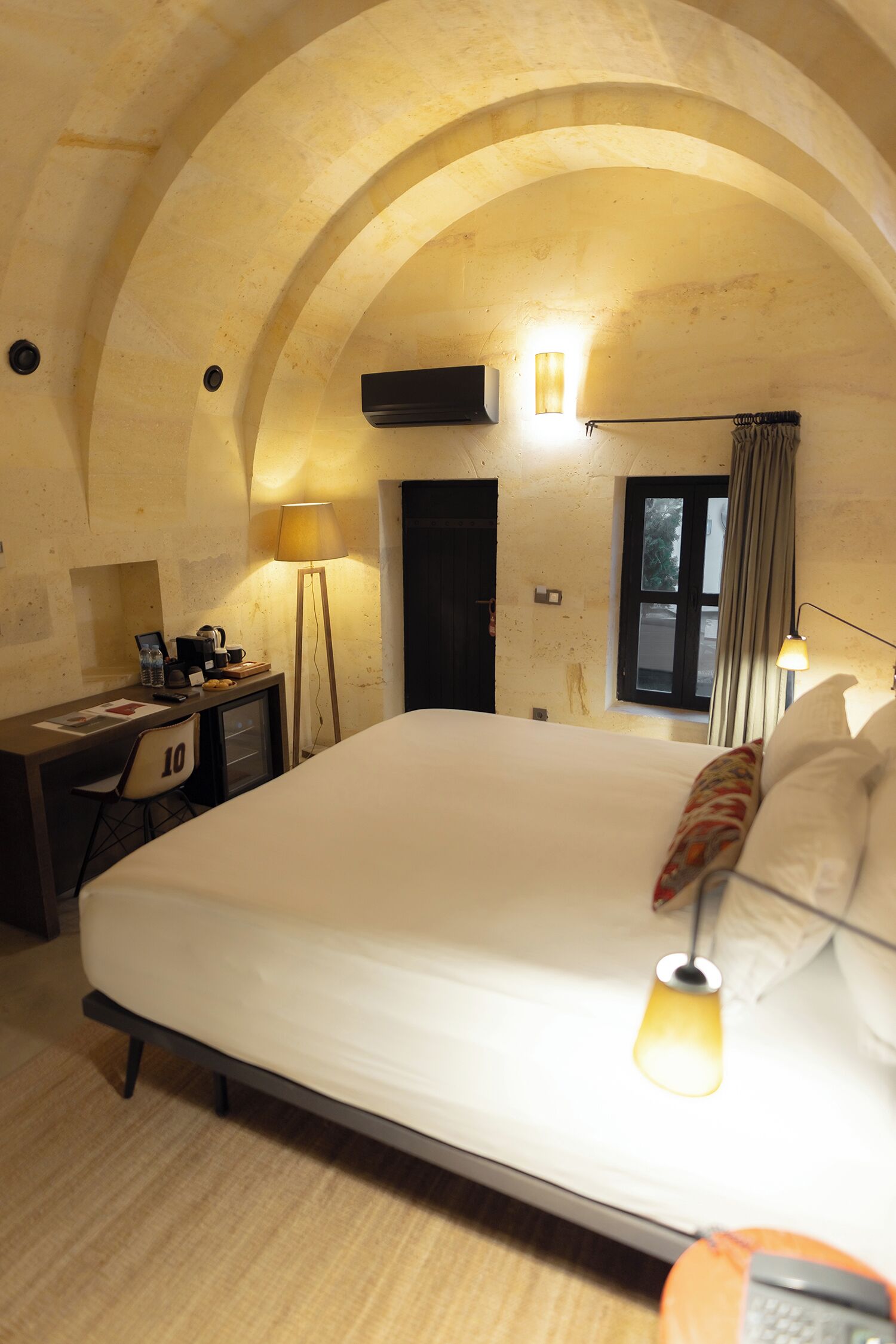 Een hotelkamer met een groot bed, bureau, stoel, een raam met gordijnen en een lamp.