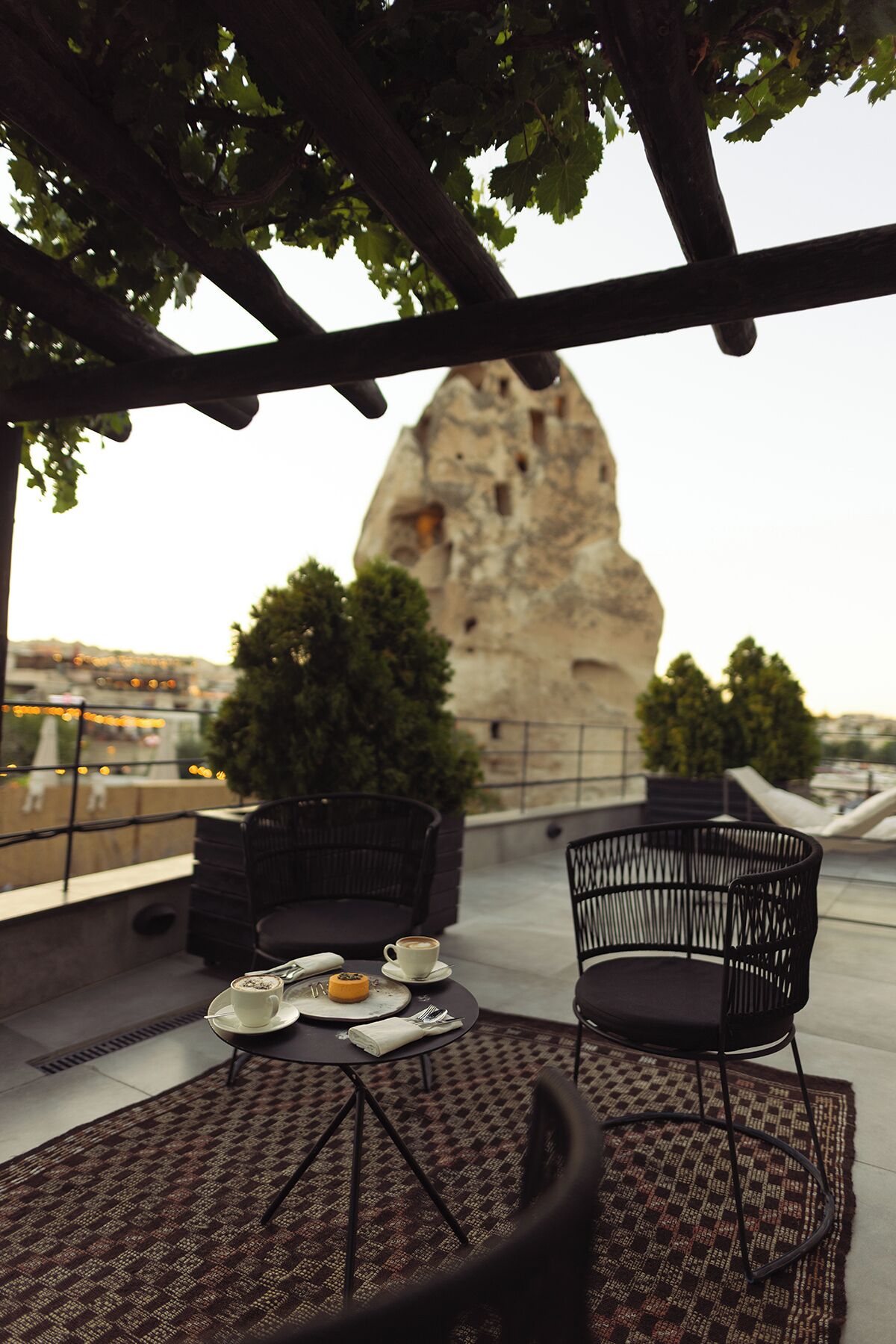 A rooftop terrace with two black wicker chairs, a table with a cup of coffee and a pastry, and a view of a large rock formation.