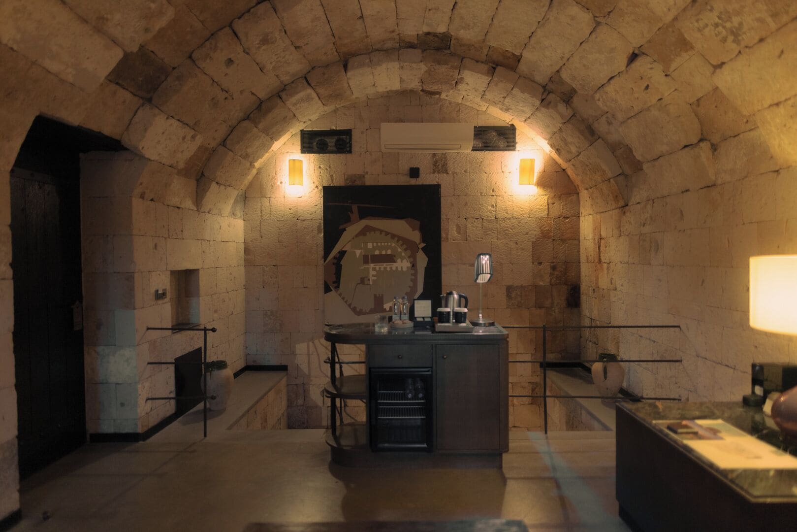 A stone-walled room with a central counter, two wall-mounted lights, and a large window.