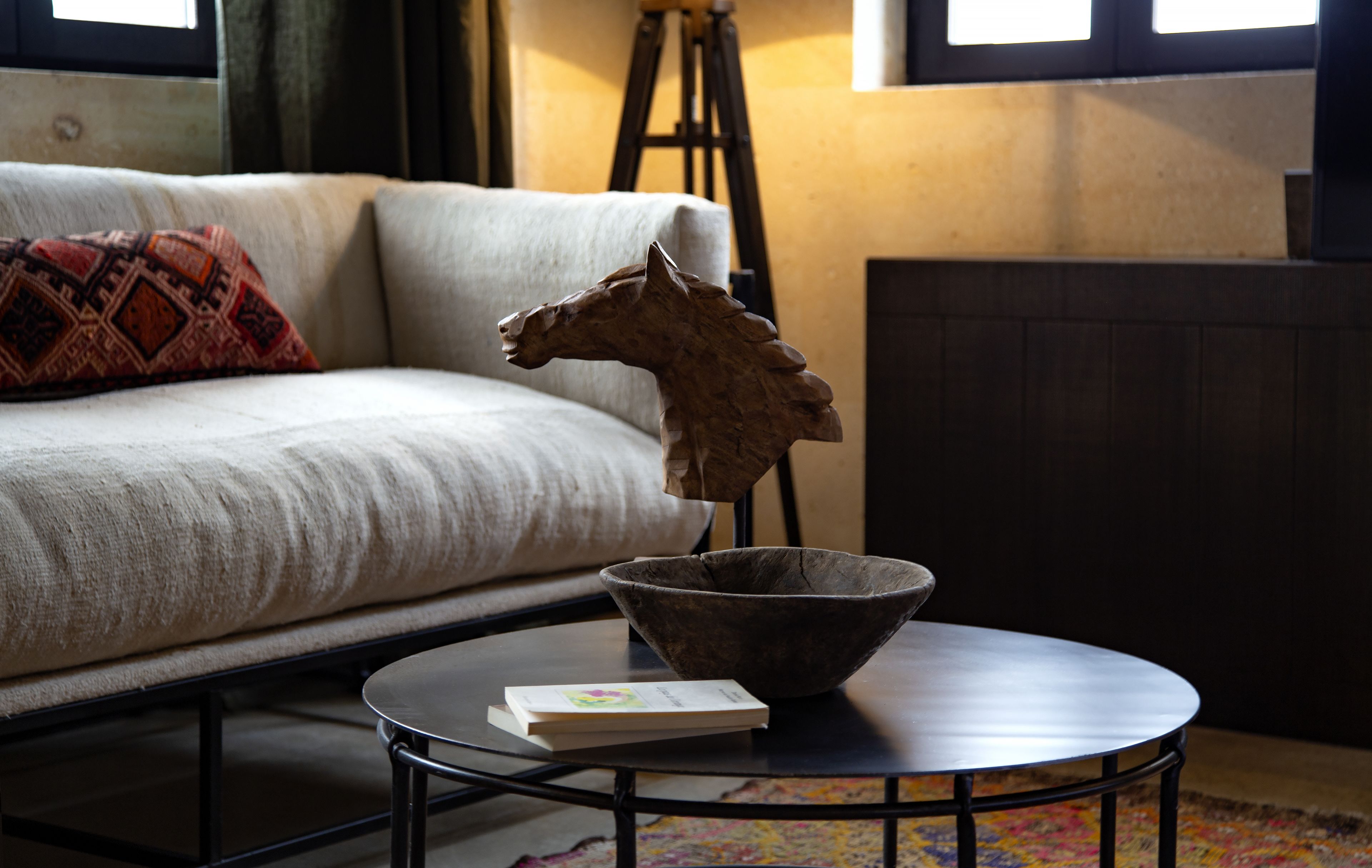 Een kamer met een bank, een klein rond tafeltje met een boek en een houten paardbeeldje, en een driepootlamp op de achtergrond.