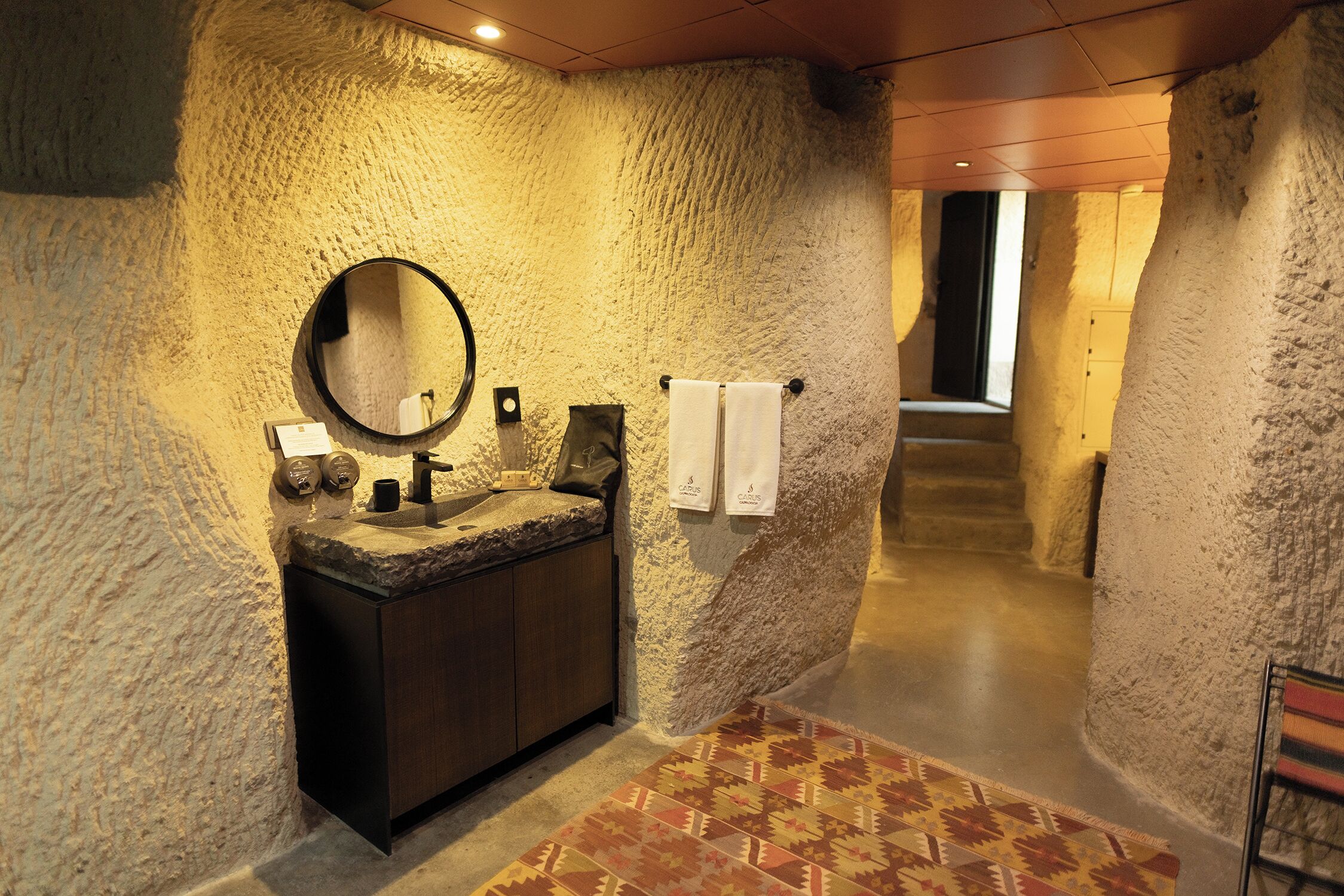 A bathroom with a stone wall, a round mirror, a sink, and a patterned rug.