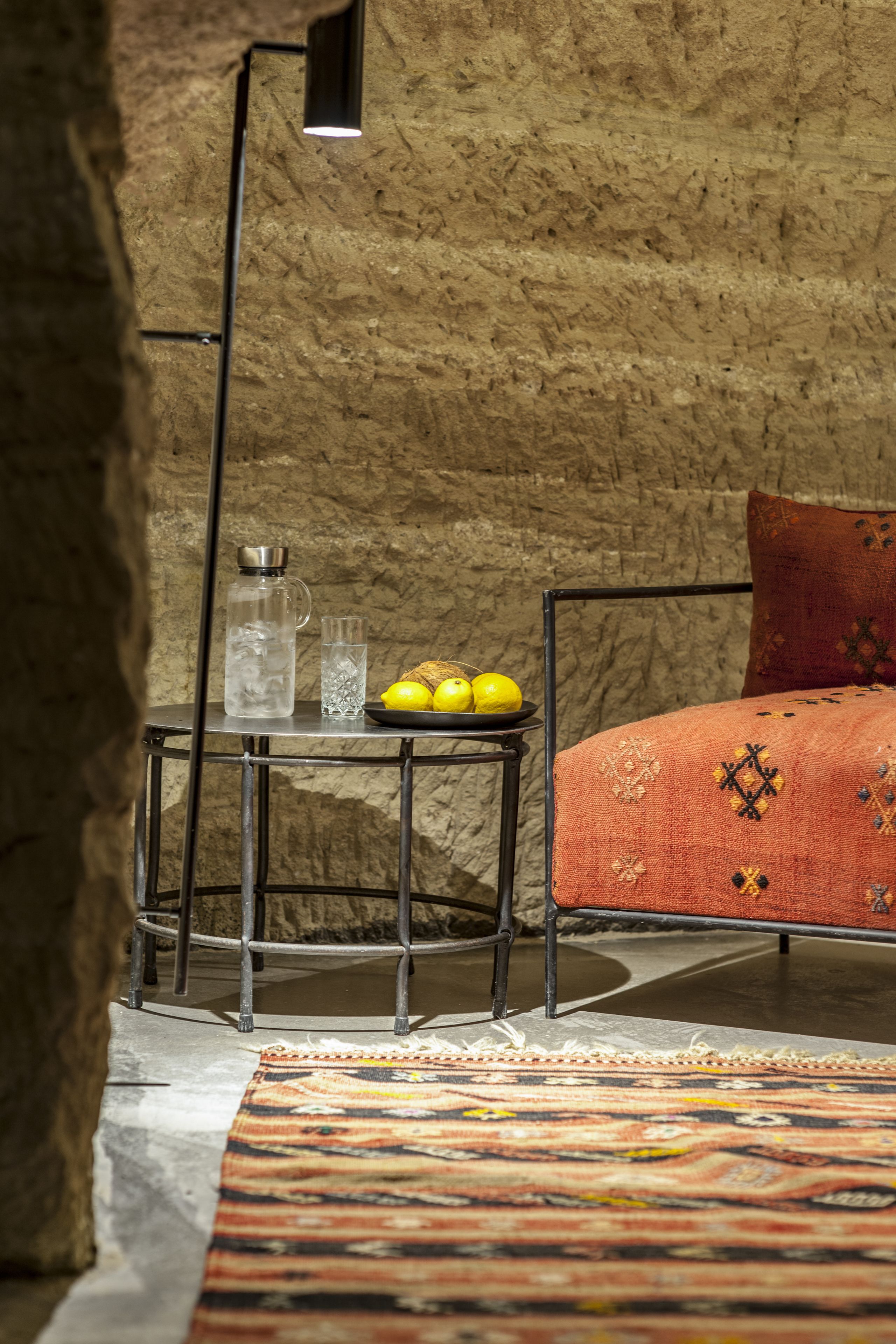A room with a rustic stone wall, a metal-framed bed with a patterned blanket, a small table with a water bottle and lemons, and a floor lamp.
