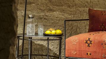 Une pièce avec un mur en pierre rustique, un lit à ossature métallique recouvert d’une couverture à motifs, une petite table avec une bouteille d’eau et des citrons, et un lampadaire.