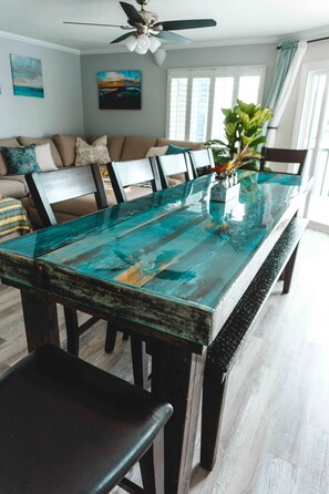 A dining table with a glass top, surrounded by chairs, in a room with a ceiling fan, a sofa, and a window with curtains.