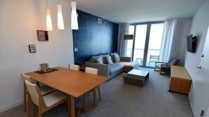 A modern living room with a wooden dining table, a sofa, and a TV mounted on the wall. - 2 Bedroom Unit Across From Ocean @ Beachwalk Resort (Hallandale Beach)