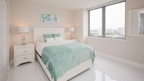 A modern bedroom with a large bed, two bedside tables, and a large window offering a city view. - Ocean View 3 Bed 3 Bath Across The Beach @ Beachwalk (Hallandale Beach)