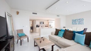 A modern living room with a sofa, armchairs, a coffee table, and a TV. A kitchen is visible in the background. - Ocean View 3 Bed 3 Bath Across The Beach @ Beachwalk (Hallandale Beach)
