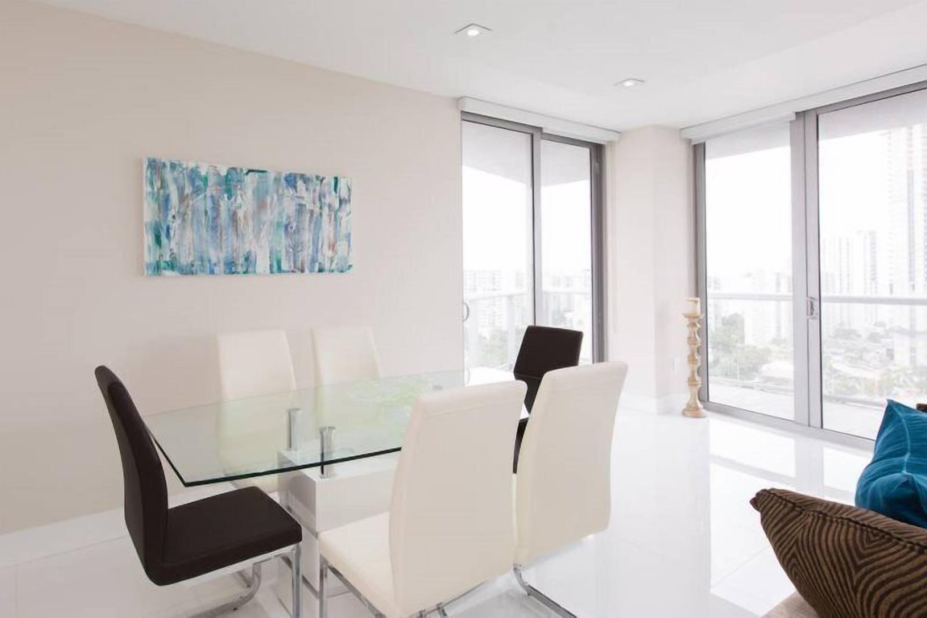 Un comedor moderno con una mesa y sillas de vidrio, vistas a la ciudad a través de grandes ventanales y una obra de arte abstracta en la pared.