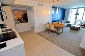 A modern kitchen with a built-in stove, a dining area with a wooden table and chairs, a living room with a sofa and a large window, and a blue accent wall. - Bay, Ocean & Skyline View 2 Bed 2 Bath Residence @ Beachwalk Resort (Hallandale Beach)