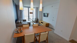 Un salon moderne comprenant un canapé, une table à manger avec des chaises, un téléviseur fixé au mur et une grande fenêtre avec des rideaux.