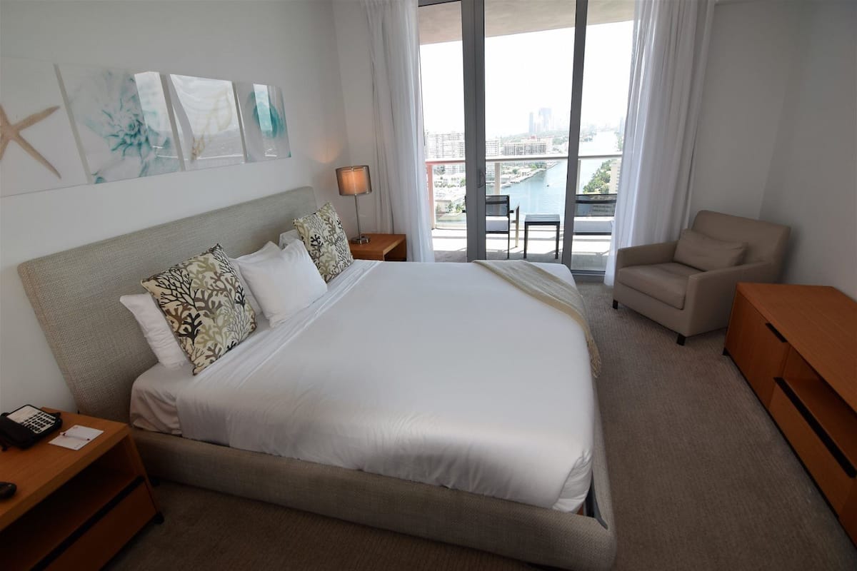 A hotel room with a large bed, a desk, a chair, and a view of the city through the window.