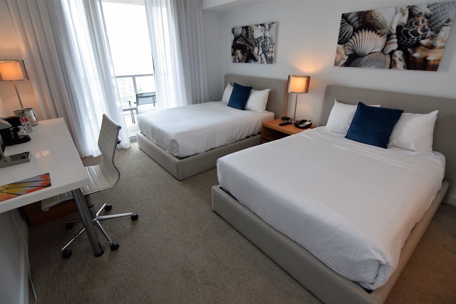 Habitación de hotel con dos camas, un escritorio y una silla. Hay dos mesitas de noche con lámparas, una ventana con cortinas y obras de arte en las paredes.