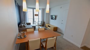 Una sala de estar moderna con un sofá, una mesa de comedor con sillas, un televisor montado en la pared y un gran ventanal con cortinas.