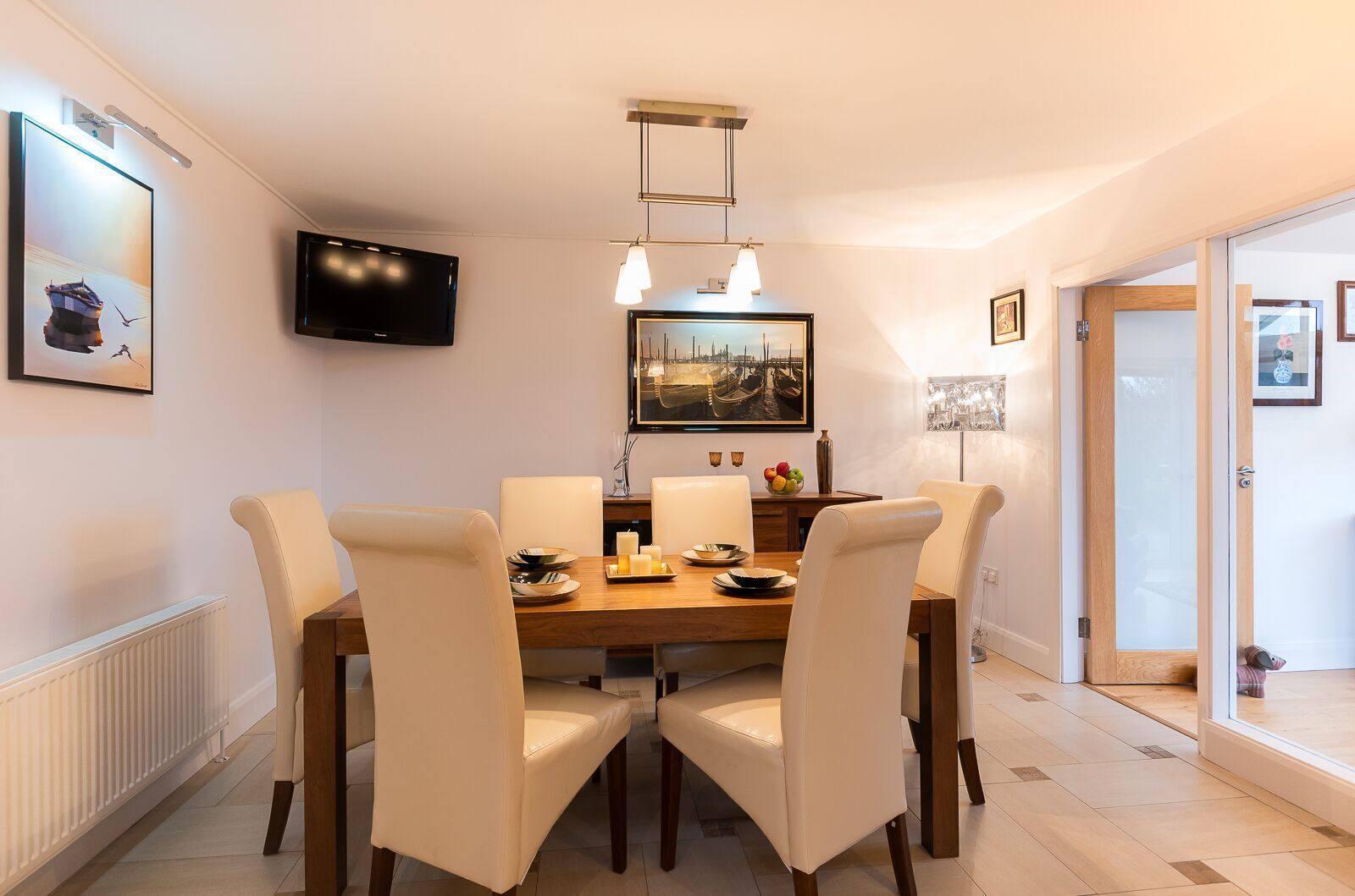 A dining area with a wooden table, four chairs, a television mounted on the wall, and a painting above the table.