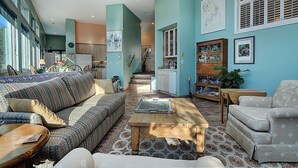 Un salón espacioso con un sofá grande, una mesa de centro y una alfombra estampada. Se observa una zona de cocina con armarios y una mesa de comedor al fondo.