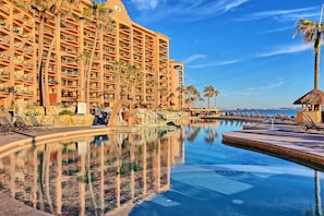 Un hôtel doté d’une grande piscine, de palmiers et surplombant un ciel bleu azur.